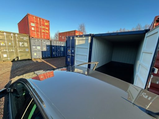 A view of stacked shipping containers, one container is open with a car in foreground at a self storage facility about to unload contents.