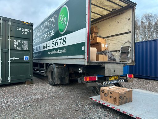 A truck with an open rear door, showing boxes and a loading ramp next to a green and blue shipping container