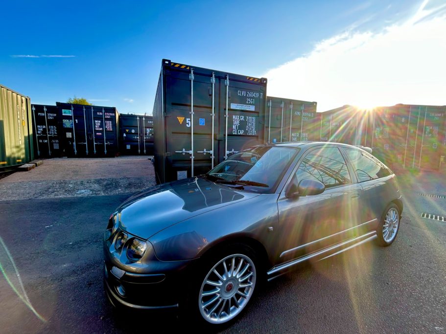 Turning circle Dark hatchback car parked near shipping containers at sunset.