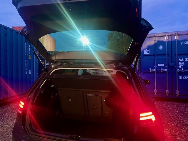 Open car boot with red taillights and a bright light in the background. Shipping containers present.