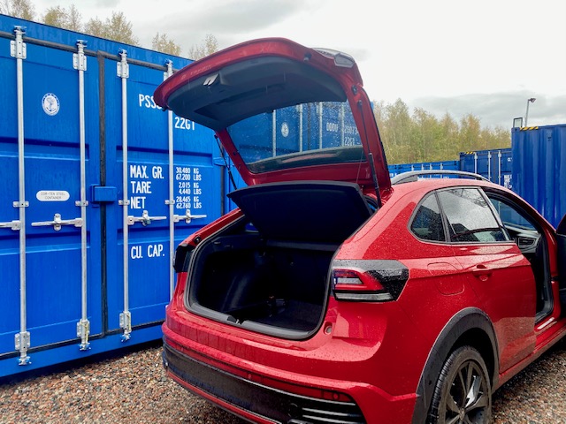 Red car with its boot open beside blue shipping containers about to put items into storage