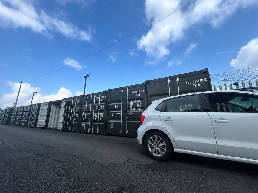 White car parked beside shipping containers under a blue sky with clouds.