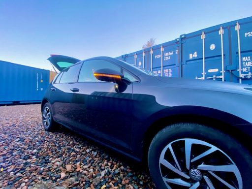 Black car with boot lid open about to unload parked beside blue shipping containers on a self storage facility.