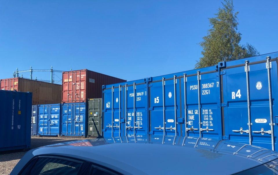 Blue shipping containers stacked in a yard under a clear blue sky.